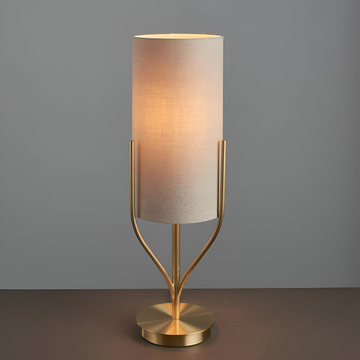 Endon Bramdon table lamp with satin brass base and slim natural linen cylinder shade, illuminated against a grey background.