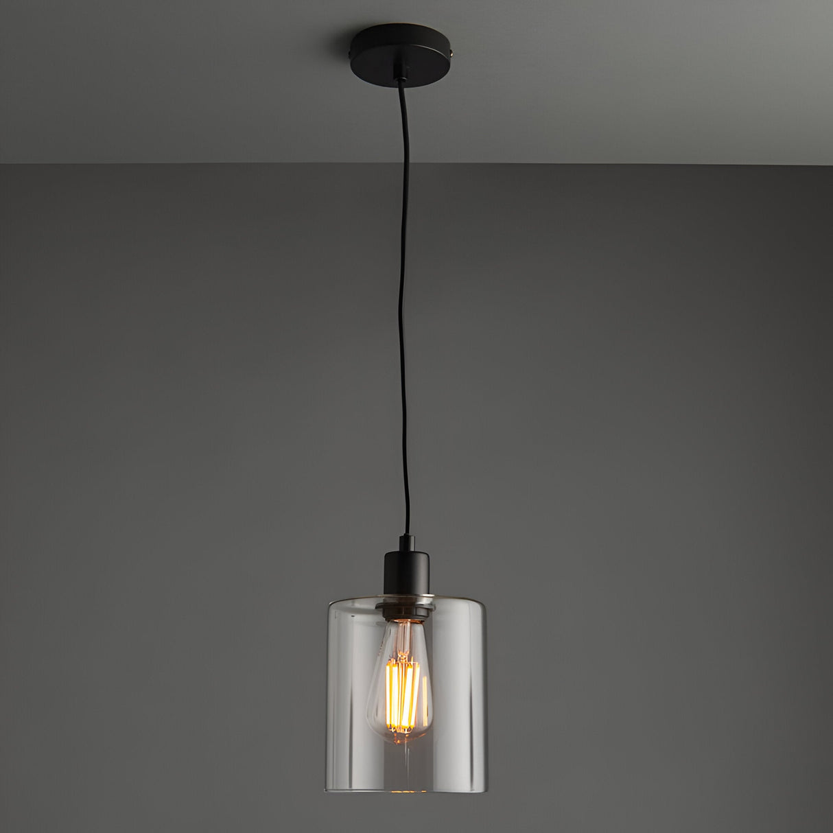 Endon Cranmere pendant light in matt black with a compact clear glass cylinder shade and exposed filament bulb on a black flex against a grey ceiling.