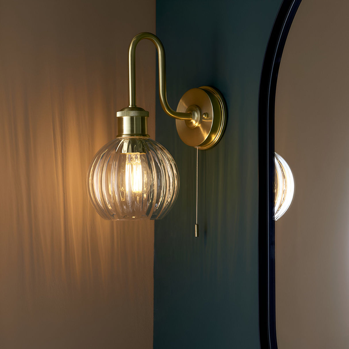 Endon Brockshaw satin brass bathroom wall light with swan-neck arm, ribbed clear glass globe shade and pull switch, mounted beside a black-framed mirror.