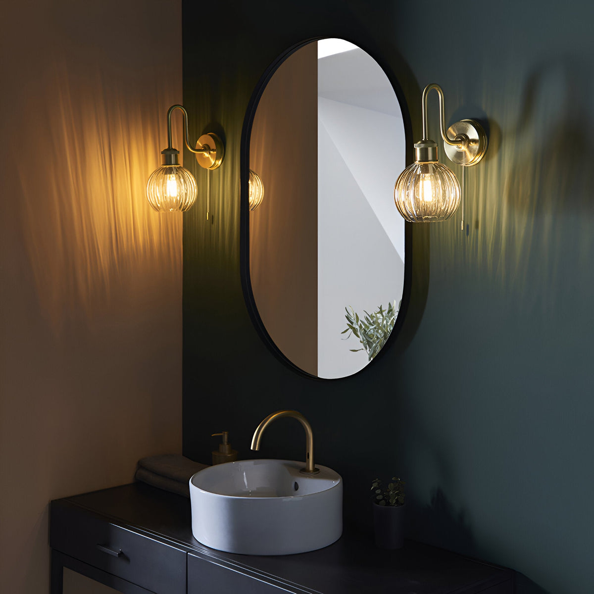 Endon Brockshaw satin brass bathroom wall lights with ribbed glass shades flanking an oval black-framed mirror above a vanity sink in a dark green bathroom.
