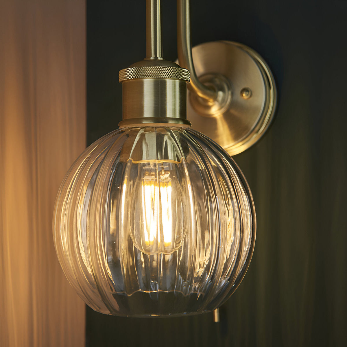 Endon Brockshaw bathroom wall light in satin brass with swan-neck arm, knurled detail and clear ribbed glass globe shade around a lit E27 bulb.