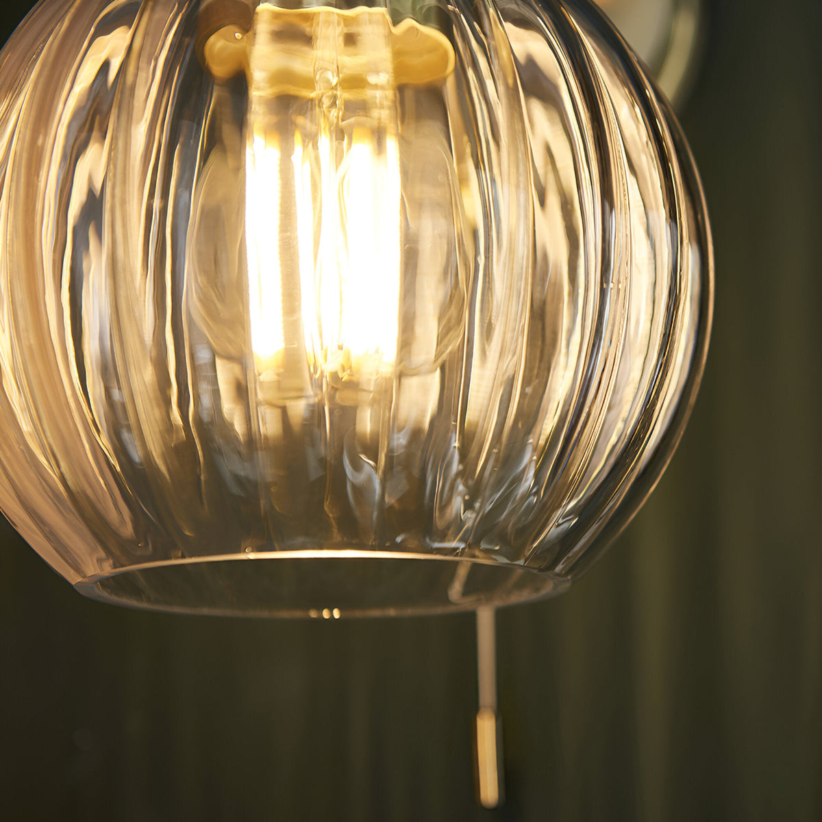 Close-up of Endon Brockshaw bathroom wall light showing clear ribbed glass globe shade illuminated by an E27 filament bulb, with satin brass pull switch below.