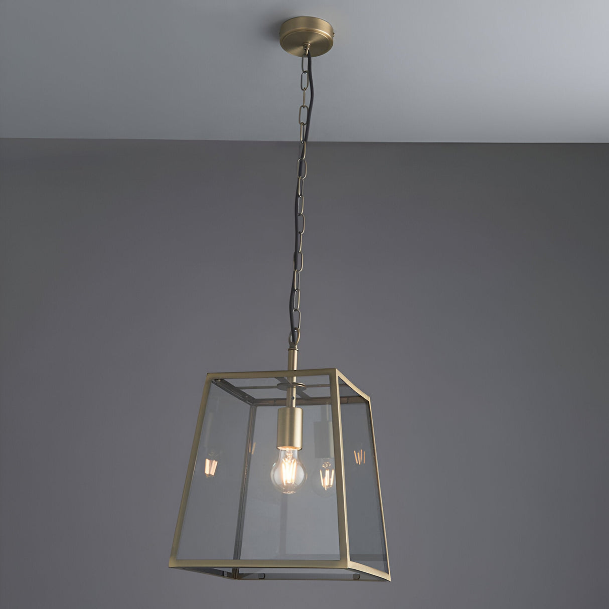 Endon Brenton lantern pendant in brass with tapered metal frame, clear glass panels and filament bulb, suspended from a chain against a grey ceiling.