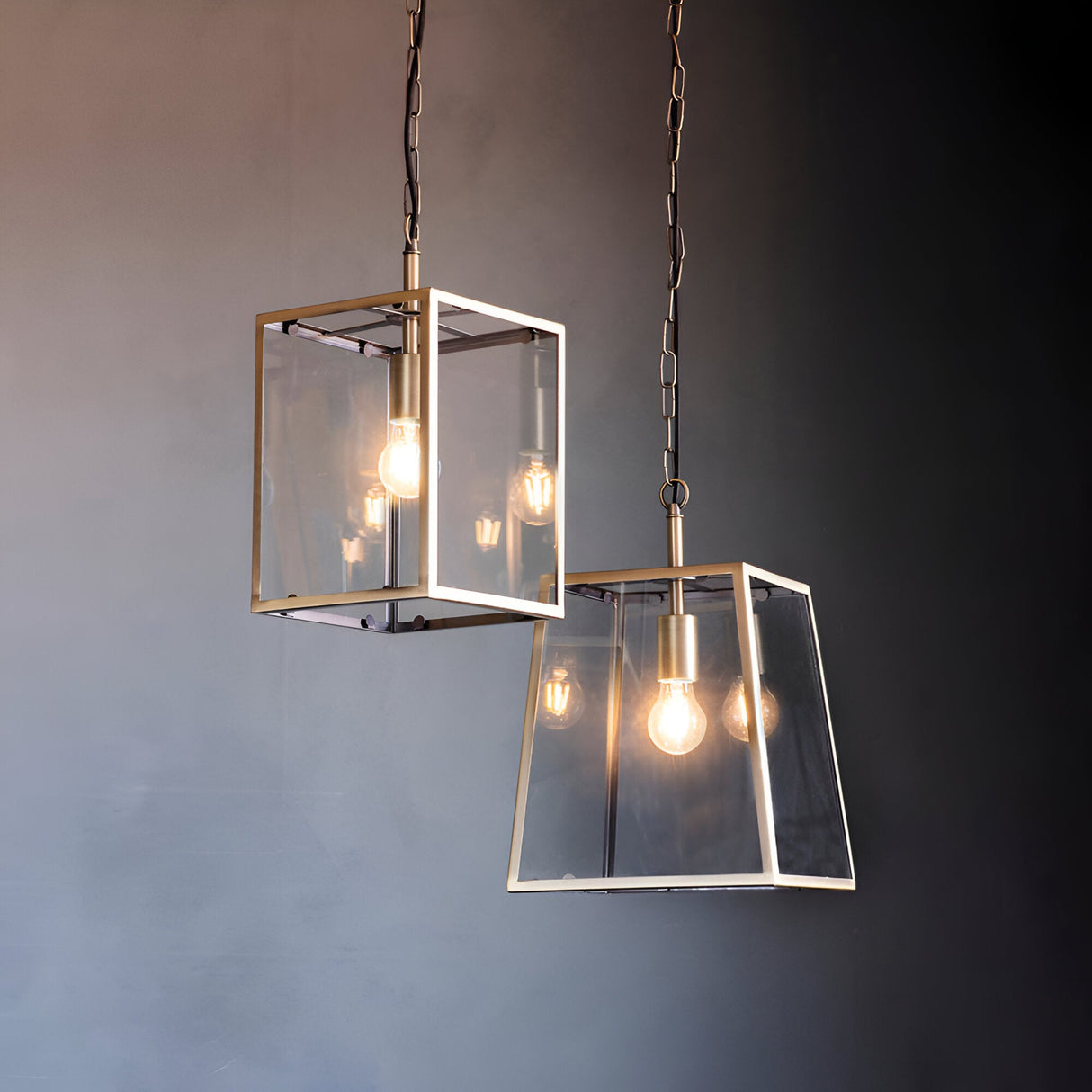 Endon Brenton lantern pendants in brass with tapered metal frames, clear glass panels, filament bulbs, and chain suspension against a dark grey wall.