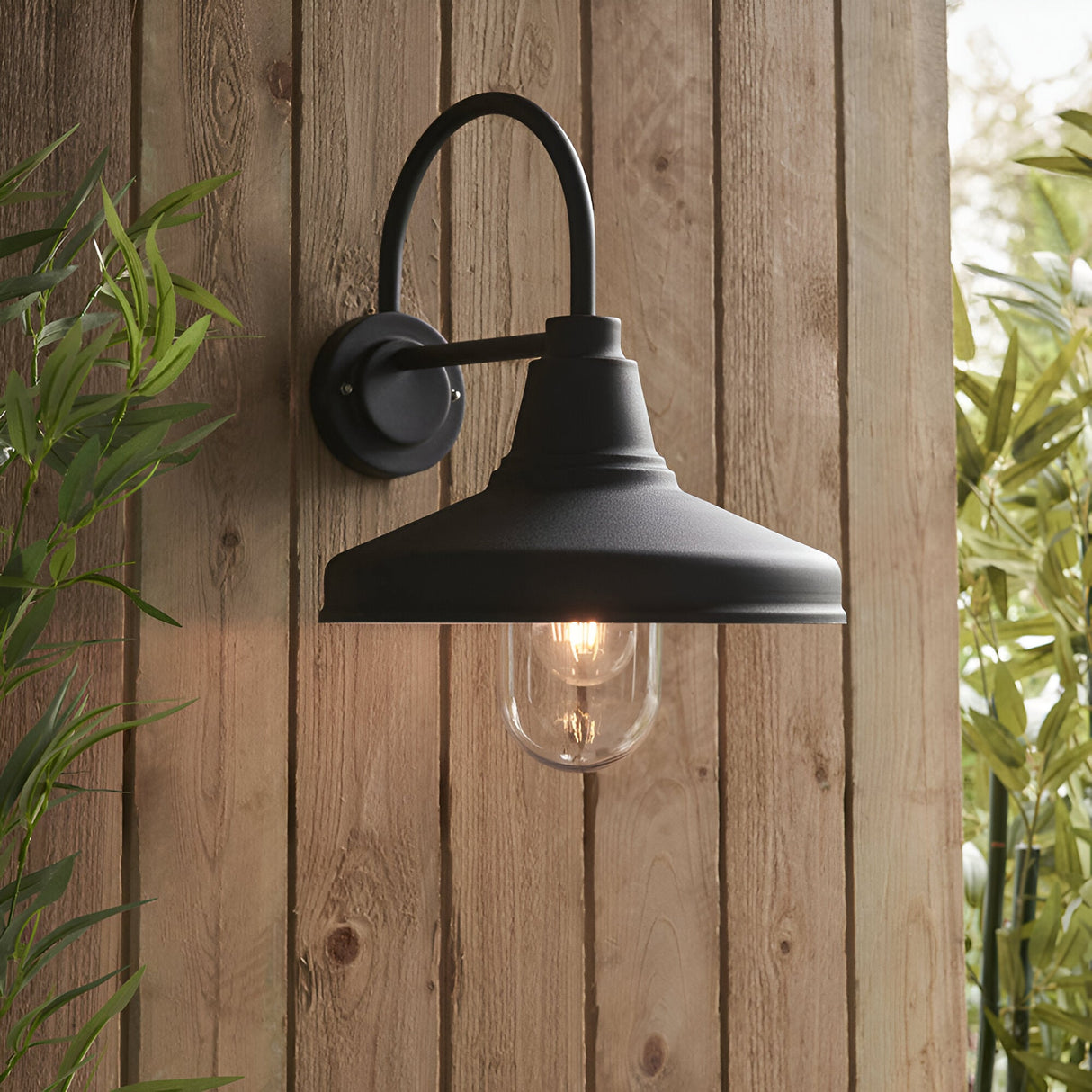 Endon Brackworth black outdoor wall light with gooseneck arm, textured metal shade and clear glass tube around an E27 filament bulb, on a wooden fence.