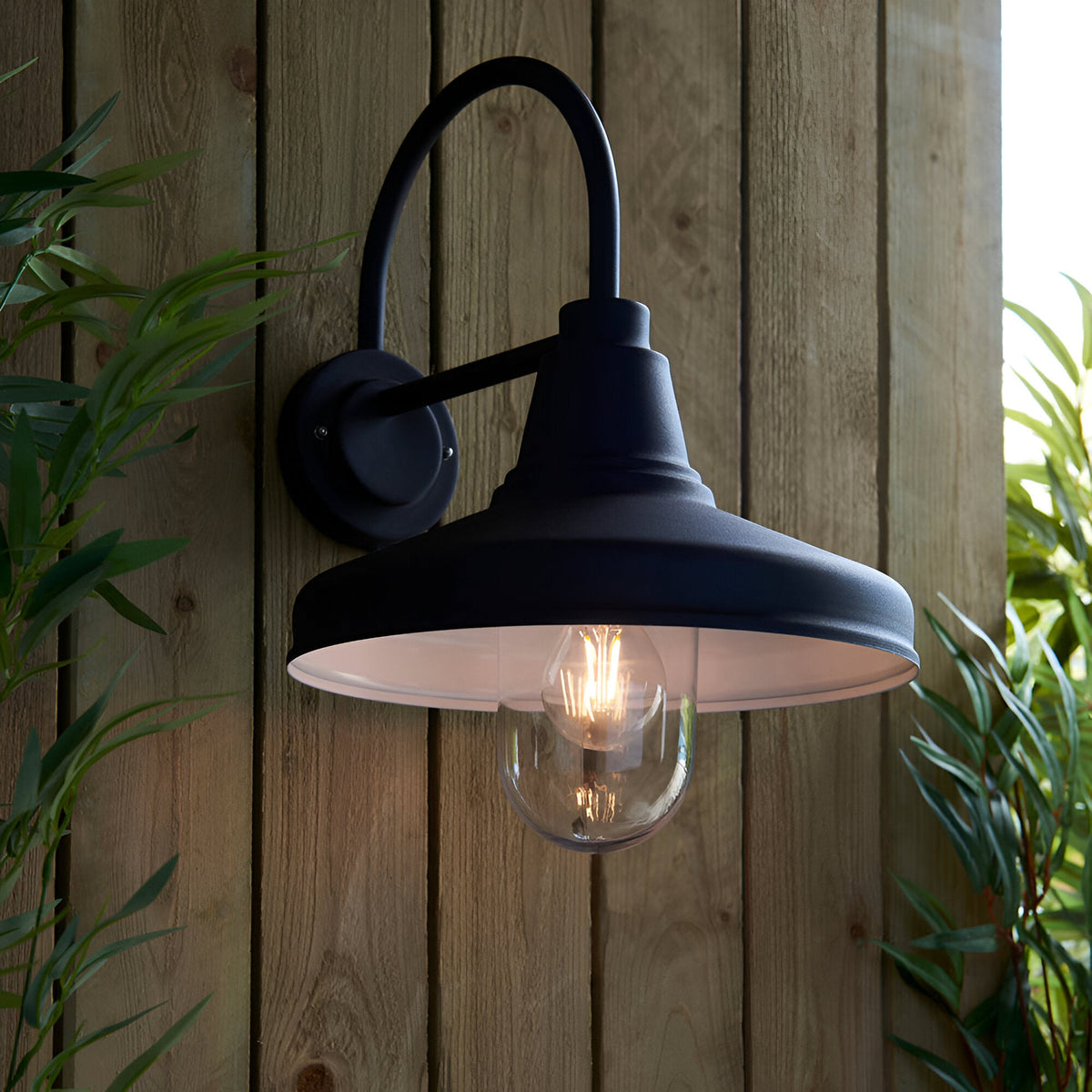 Endon Brackworth outdoor wall light in textured black with a gooseneck arm, barn-style shade and clear glass globe housing a filament bulb, on a wooden fence.