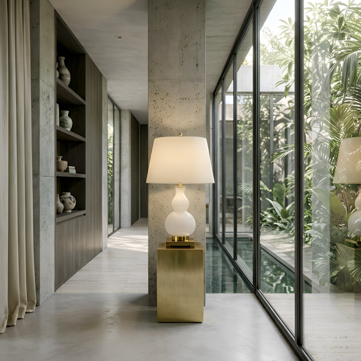 A modern interior showcases the Lumetra 1 Light Sculpted Alabaster Table Lamp in Brass & Soft White on a gold pedestal by tall windows with views of greenery and a pool, while built-in shelves on the left display decorative vases.