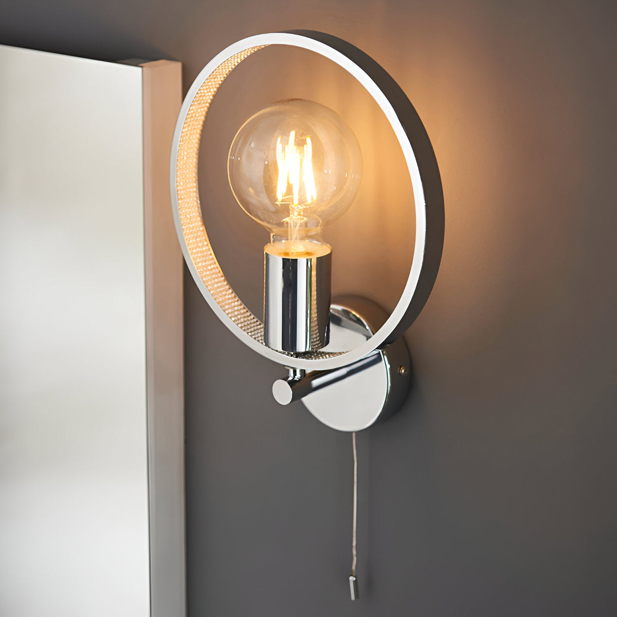 Endon Carlcombe chrome bathroom wall light with a faceted circular ring frame, single E27 globe bulb and pull cord switch, mounted beside a mirror.