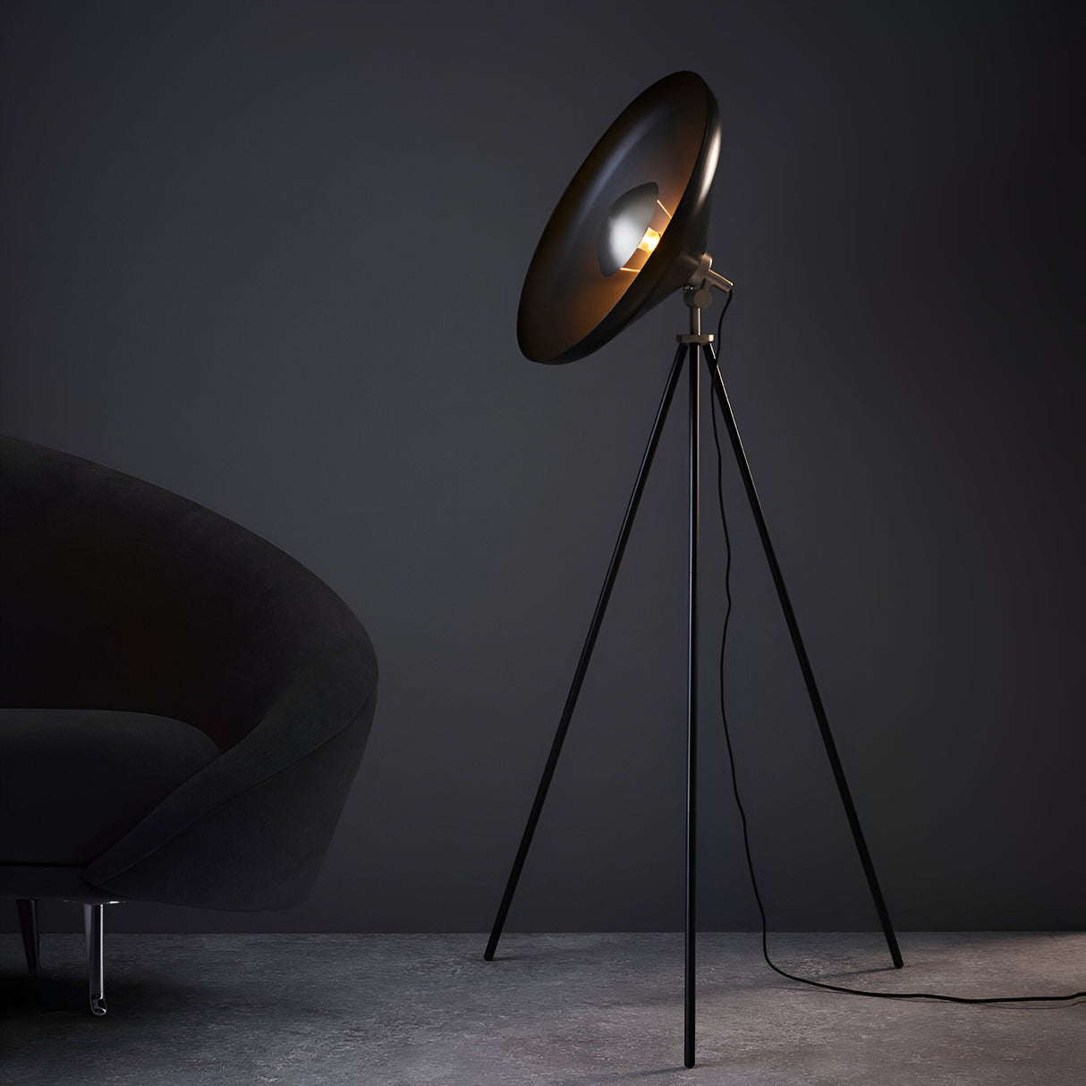 Endon Bramshaw tripod floor lamp in matt black with nickel details and large angled dome shade, beside a dark velvet armchair against a grey wall.