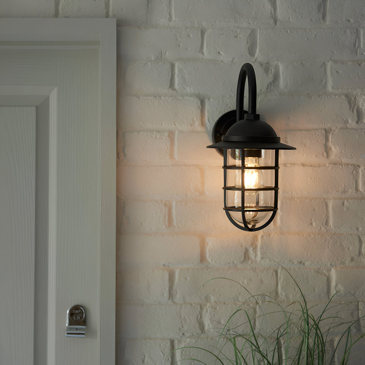 Chartgrove Outdoor Wall Light in black aluminium with caged metalwork over a clear glass shade, mounted on a whitewashed brick wall beside a door.