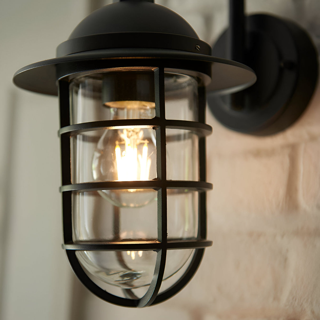 Chartgrove Outdoor Wall Light in black, close-up of caged metalwork over clear glass shade with lit E27 bulb, mounted on brick wall.