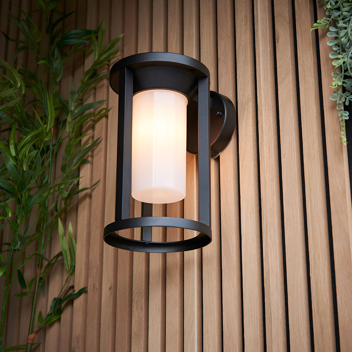 Endon Ashdale outdoor wall light with a cylindrical white polycarbonate shade in a matte black cage frame, mounted on a slatted wood wall beside foliage.