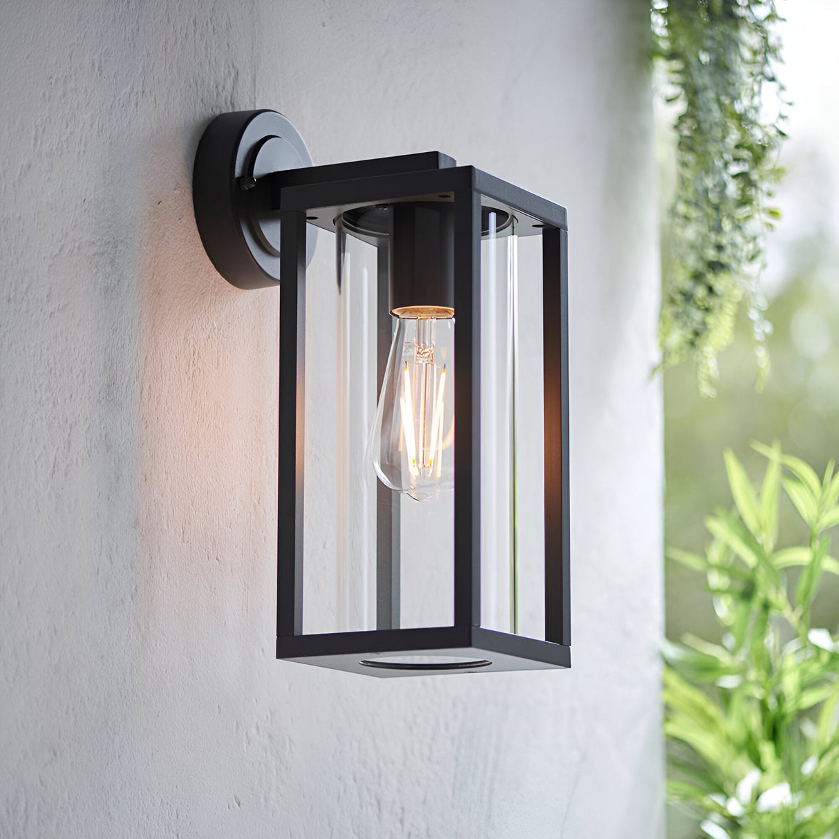 Endon Bramwood black outdoor wall light with slim rectangular lantern frame and clear glass panels, mounted on a white exterior wall beside trailing greenery.