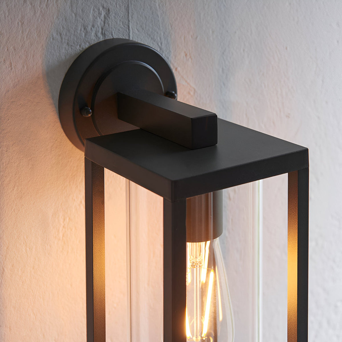 Endon Bramwood outdoor wall light in textured black aluminium, close-up of the round wall plate, slim arm and clear glass lantern with filament bulb.