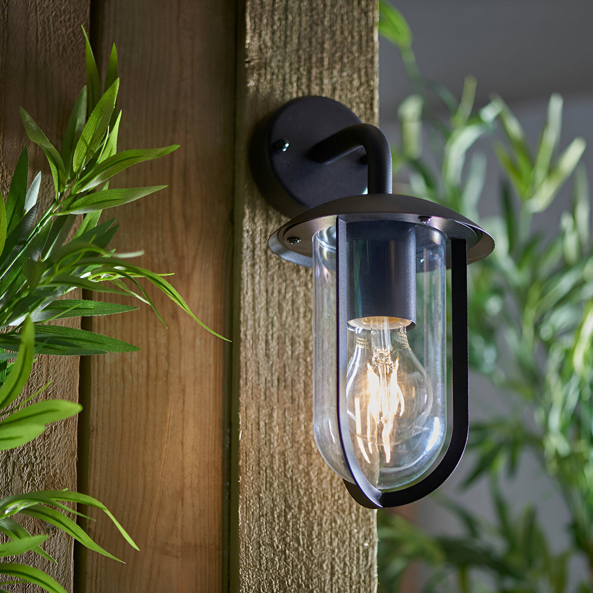 Chartley Outdoor Wall Light in black, aluminium wall bracket with clear shade and E27 filament bulb, mounted on a wooden post amid greenery.