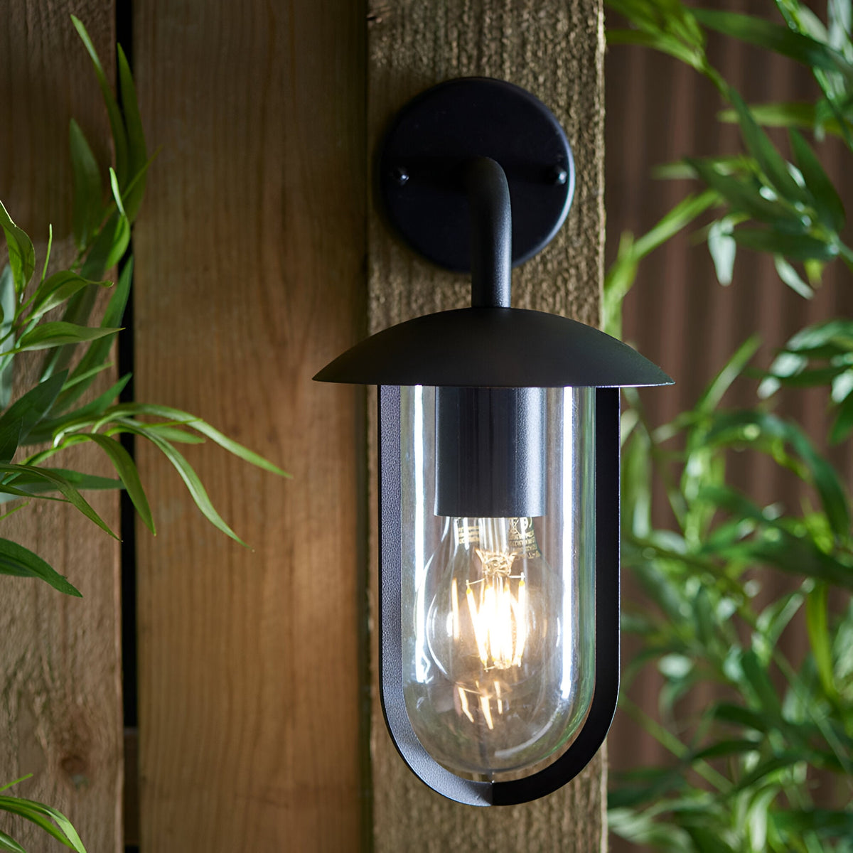 Chartley Outdoor Wall Light in black textured aluminium, mounted on wooden fence with a clear shade housing a lit E27 filament bulb, surrounded by greenery.