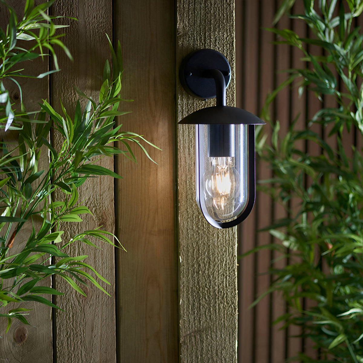 Chartley Outdoor Wall Light in black aluminium, mounted on a wooden fence panel with a clear shade revealing a glowing filament bulb, framed by green foliage.