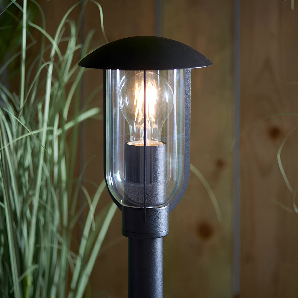 Chartley Outdoor Post Light in textured black aluminium with clear polycarbonate shade, lit E27 bulb visible, set against a wooden fence and foliage.