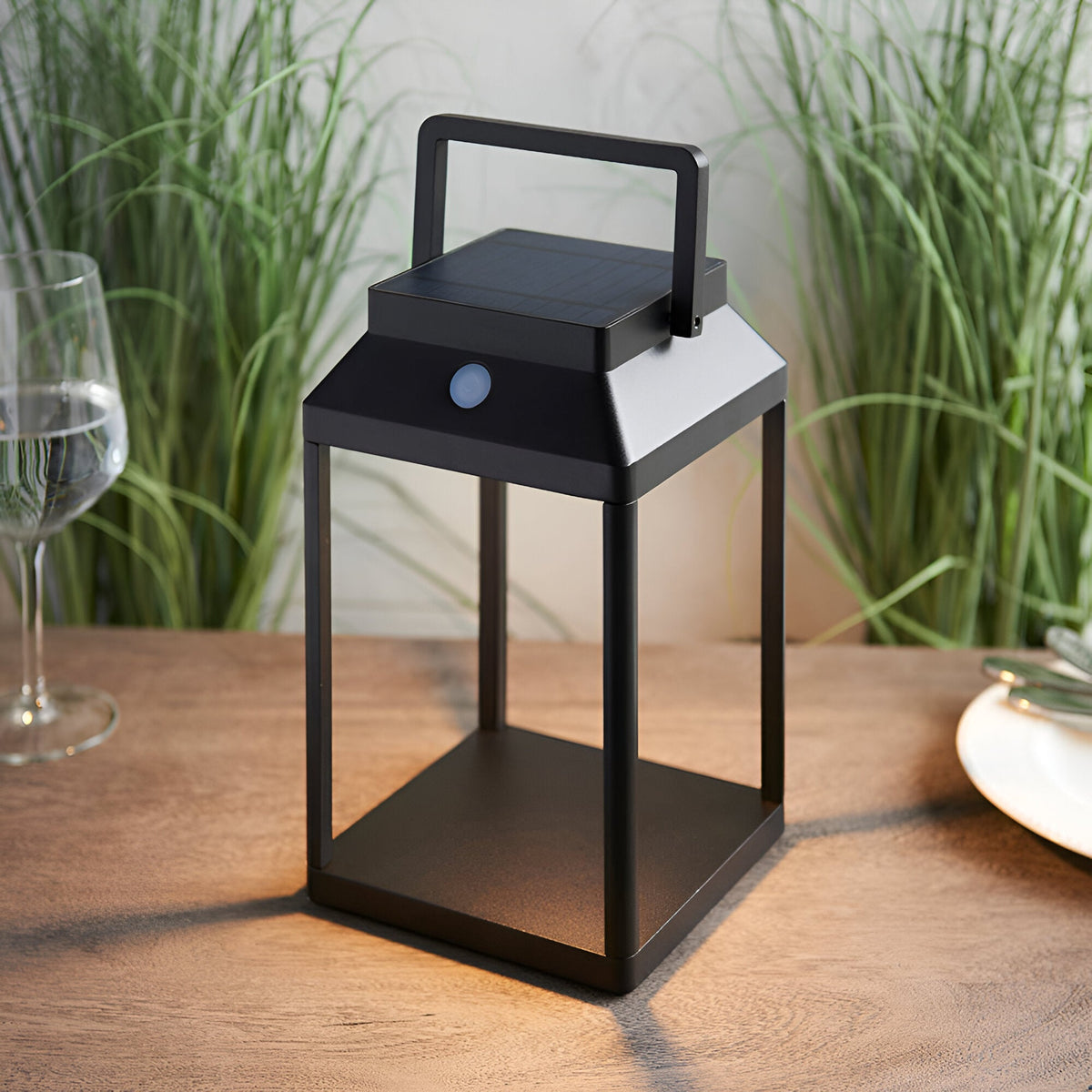Caldshaw LED Solar Table Lamp in black, lantern-style aluminium frame with white diffuser panel and carry handle, shown lit on a wooden table.