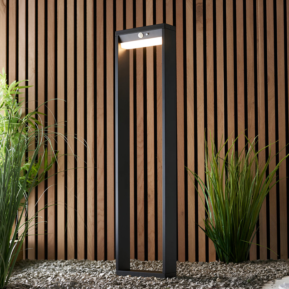 Endon Blythe LED solar bollard light in textured black aluminium with white diffuser and PIR sensor, set on gravel against a wooden slat wall with grasses.