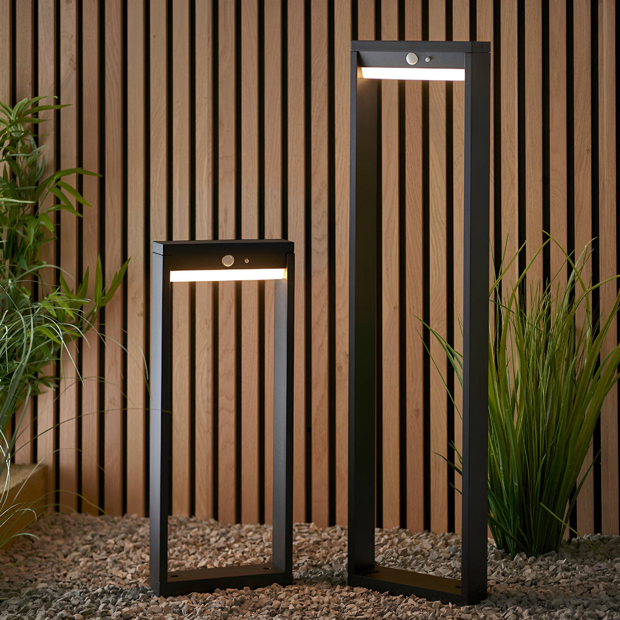 Endon Blythe LED solar bollard lights in black aluminium with PIR sensors and warm white diffusers, in tall and short heights on gravel by a wood wall.