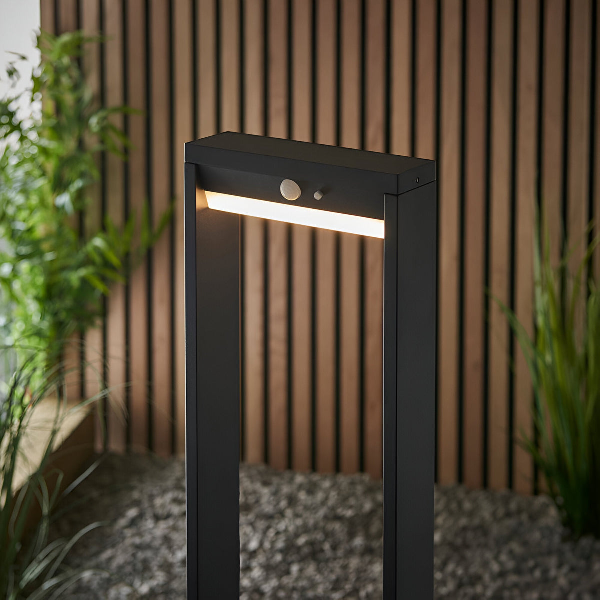 Endon Blythe LED solar bollard light in black aluminium with warm white diffuser and PIR sensor, shown on gravel against a slatted wood wall.