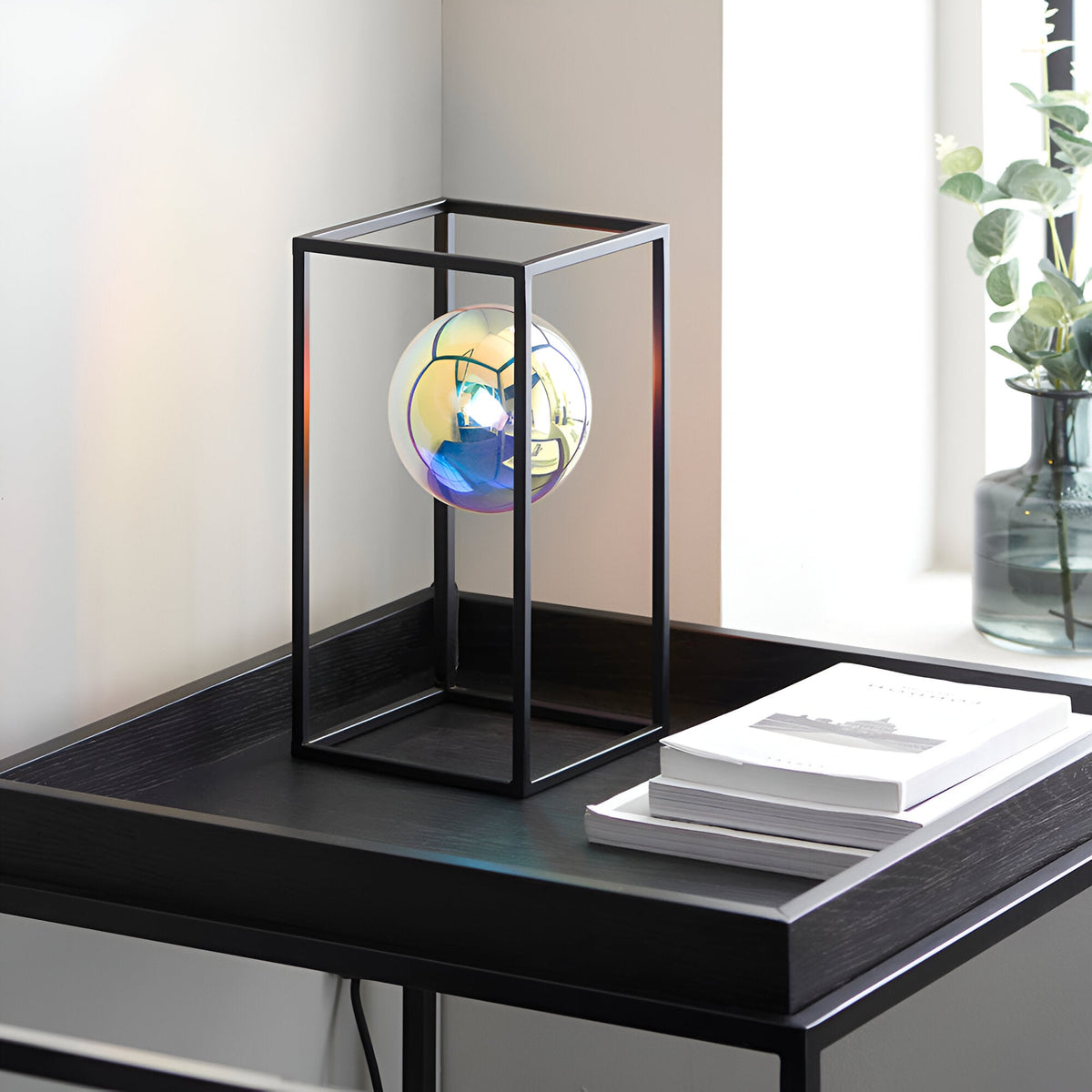Brockwell Table Lamp in matt black, open rectangular frame with iridescent orb glass shade, shown on a bedside table beside books and a vase.