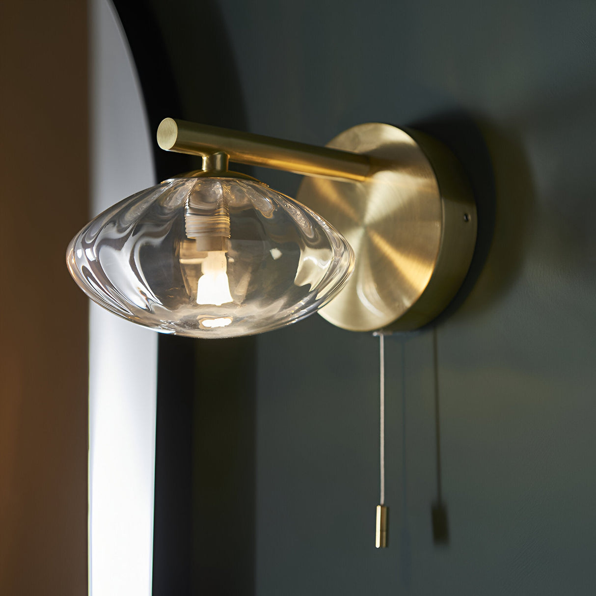 Carlmont Bathroom Wall Light in satin brass, single arm with ribbed glass shade and pull cord switch, mounted on a dark wall.