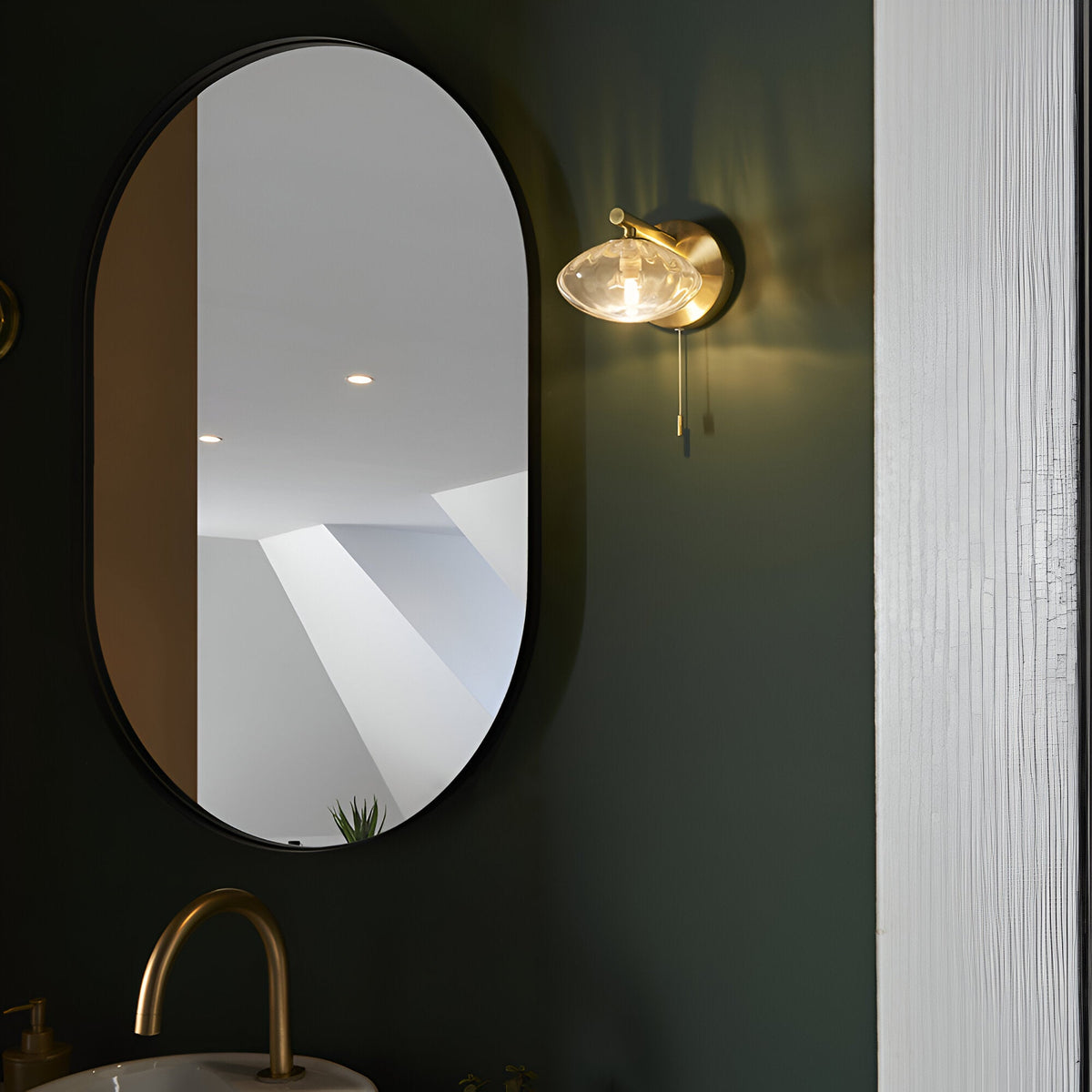 Carlmont Bathroom Wall Light in satin brass with ribbed glass shade and pull switch, shown beside an oval mirror on a dark green bathroom wall.