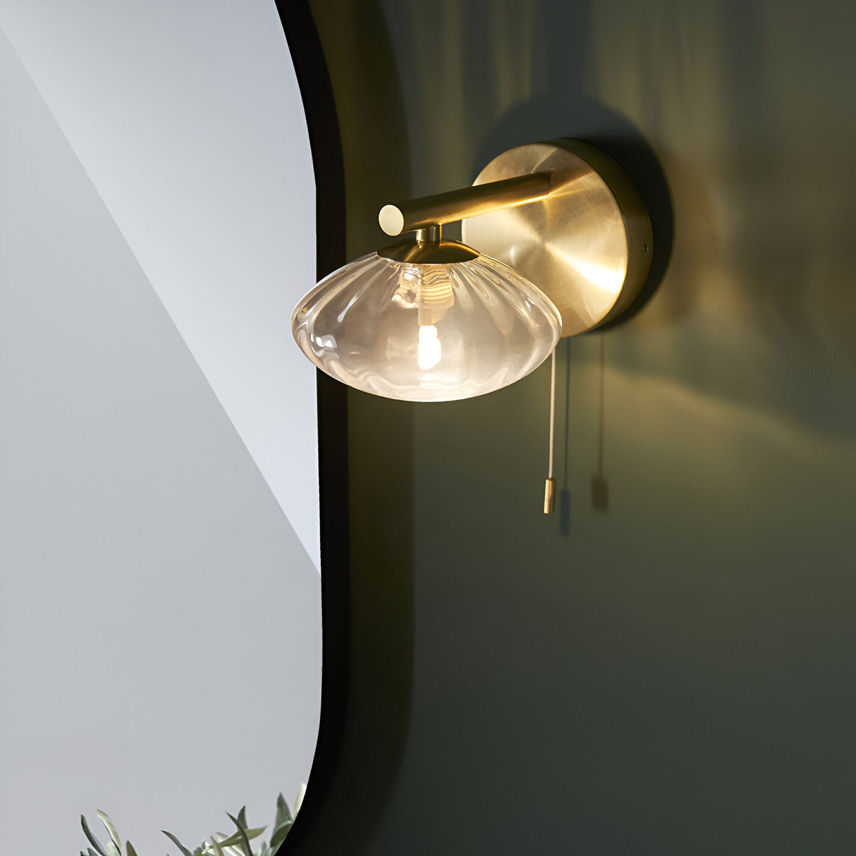 Carlmont Bathroom Wall Light in satin brass, with a rod arm holding a ribbed glass shade and pull cord switch, shown beside a mirror.