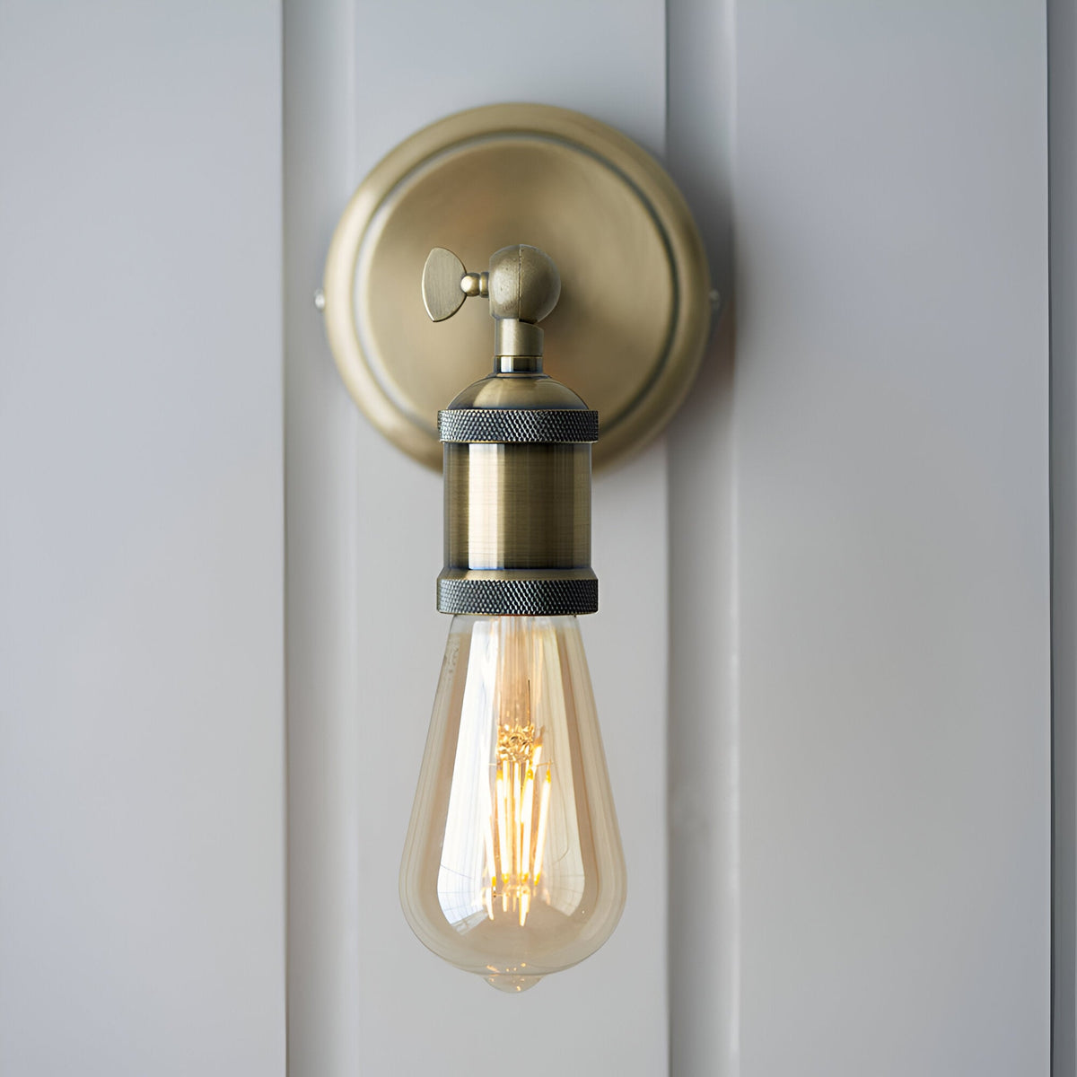 Antique brass Brackley wall light, industrial-style single E27 fixture with round backplate, adjustable knurled head, mounted on a white panelled wall.