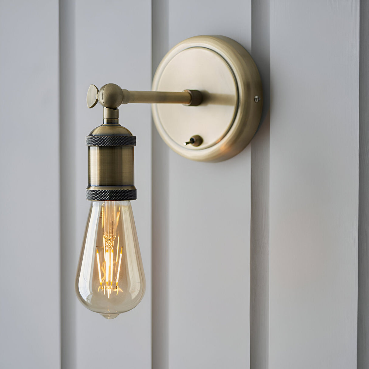 Brackley Wall Light in antique brass, industrial-style single-light fitting with adjustable arm and exposed E27 bulb, mounted on light grey panelled wall.