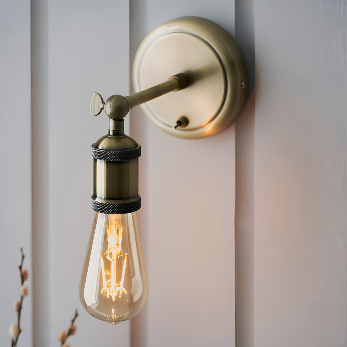 Antique brass Brackley wall light with adjustable arm, knurled knob, and E27 fitting, shown lit with an exposed filament bulb.