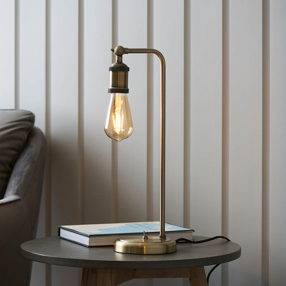 Brackley Table Lamp in brass with adjustable knuckle joint and exposed E27 bulb, shown on a side table beside a book in a living room setting.