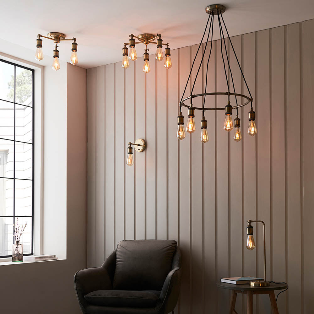 Brackley Table Lamp in brass, adjustable industrial-style design with knuckle joint and exposed bulb, shown on a side table beside an armchair in a living room.