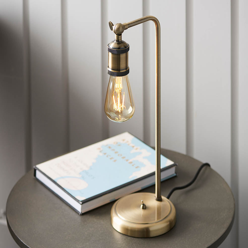 Brackley Table Lamp in brass with an adjustable industrial-style arm and exposed bulb, shown beside a book on a round side table.
