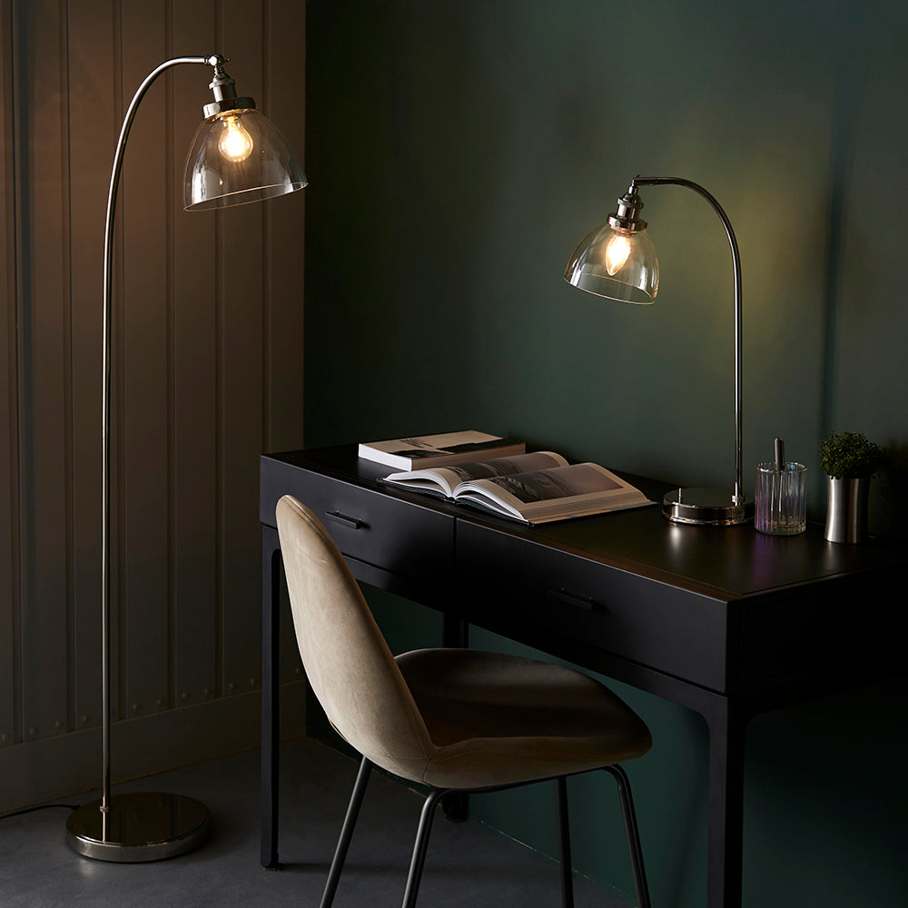 Endon Wentworth nickel floor lamp with arched stem and clear glass dome shade, lit beside a black desk with matching table lamp in a dark green room.
