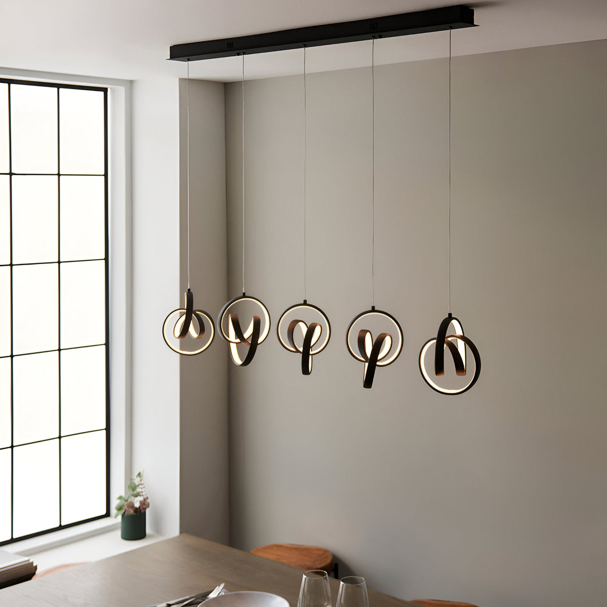 Bevwell 5 Light Linear Pendant in matt black metal, looped ring designs with integrated LEDs, suspended over a dining table in a neutral-toned room.