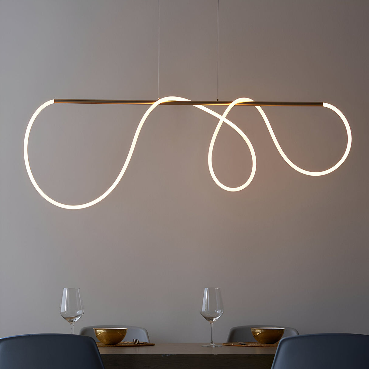 Aldley Linear Pendant in satin gold with a flexible LED tube curved into loops, suspended over a dining table with glassware and chairs below.