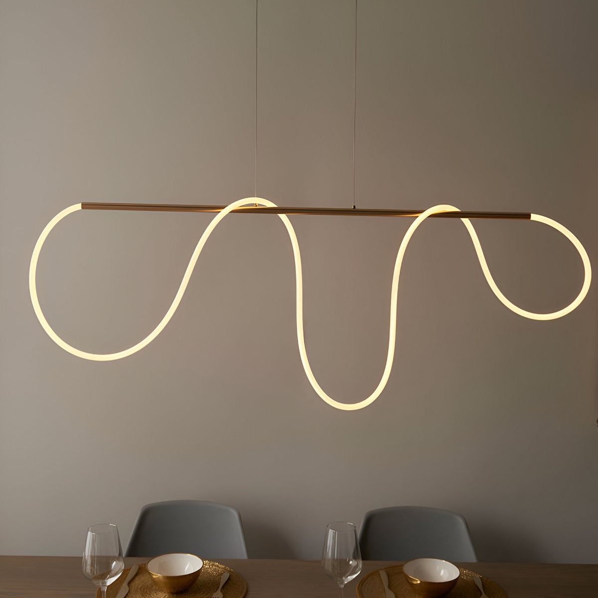 Aldley Linear Pendant in satin gold, a horizontal bar with a flexible LED tube curving into wave shapes, hung above a dining table with grey chairs.