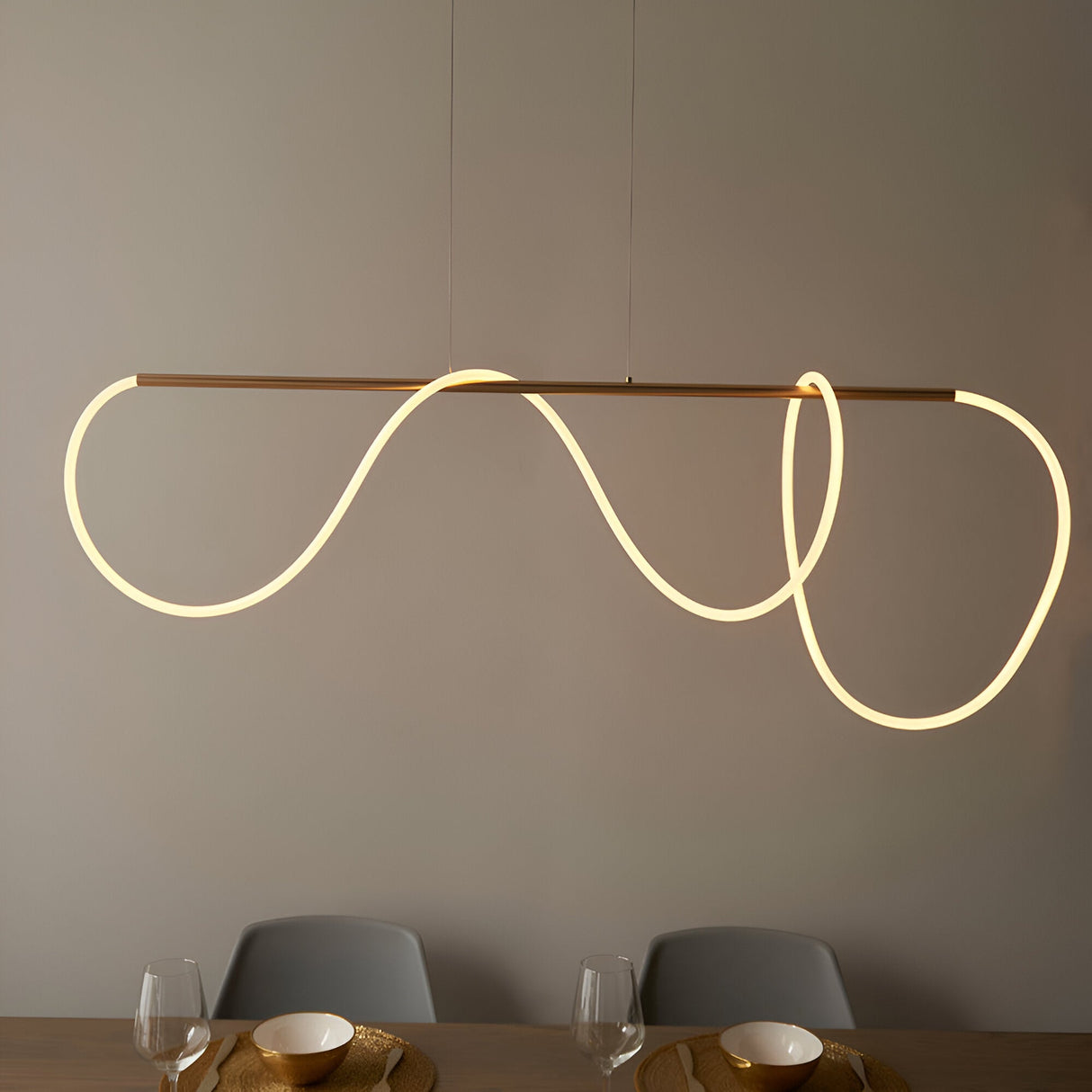 Aldley Linear Pendant in satin gold, a straight bar with a flexible warm-lit tube looping into curved waves, hung above a dining table with grey chairs.