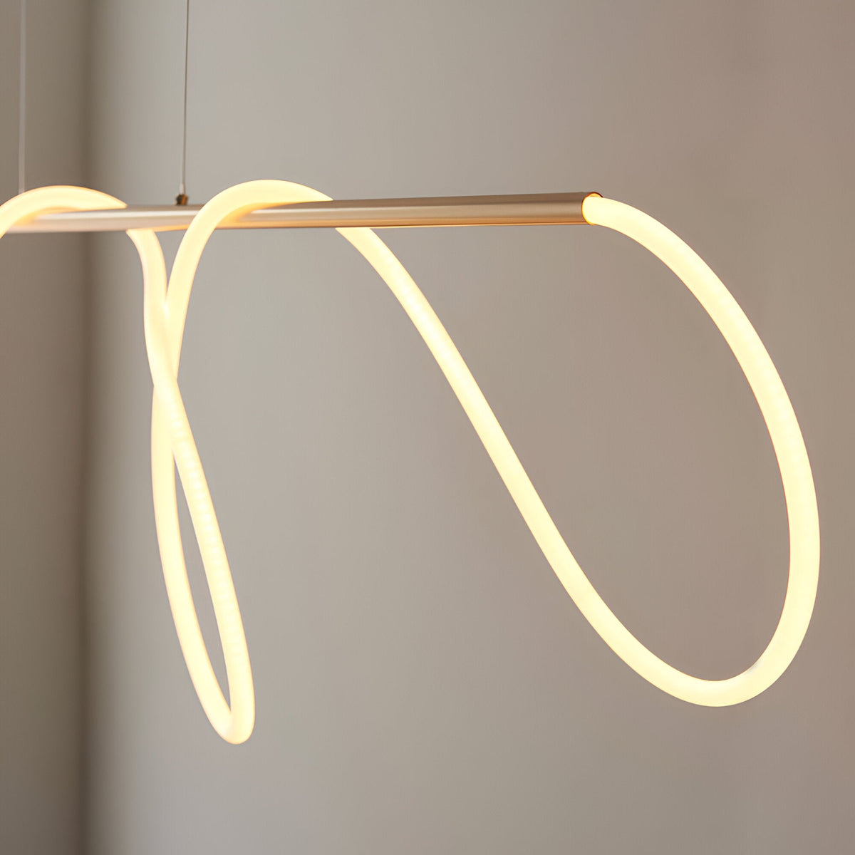 Aldley Linear Pendant in satin gold, close-up of the metal bar with a flexible warm-lit LED tube looping into soft curves against a plain grey background.