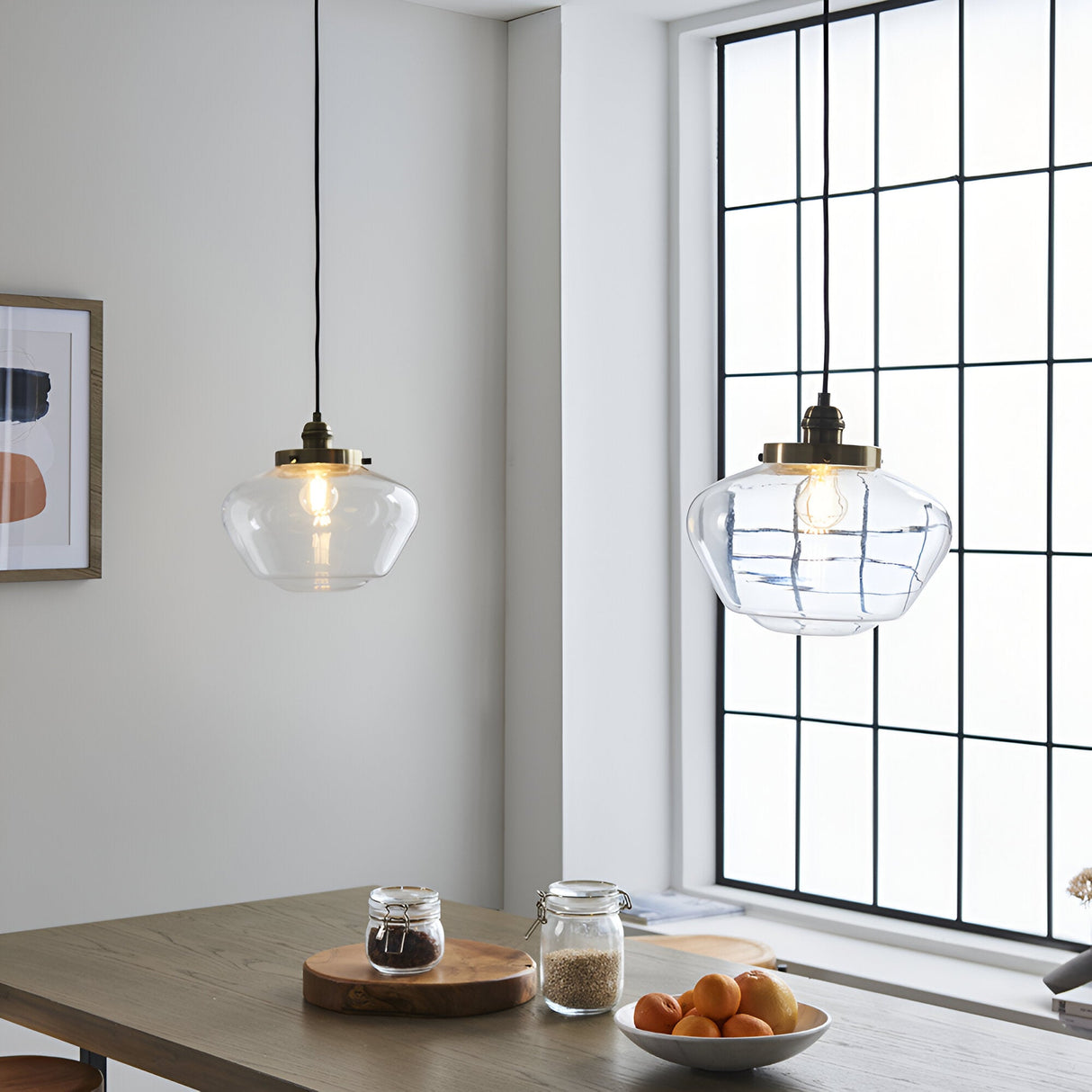 Kedleston Pendant in brass and clear glass with a schoolhouse-shaped shade, hanging above a dining table near a window in a bright kitchen.