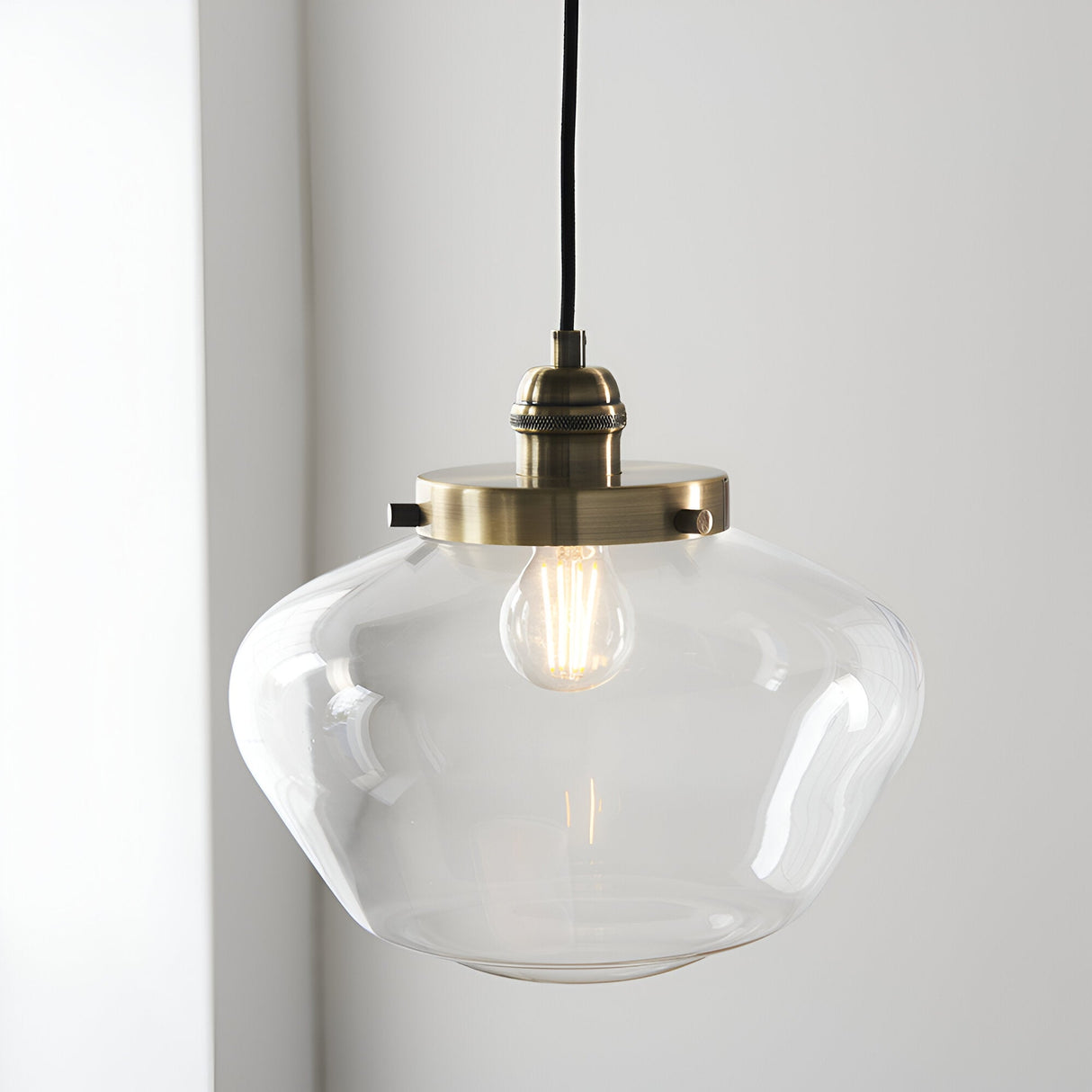 Kedleston Pendant in brass with a clear rounded glass shade and exposed E27 bulb, hung against a plain light grey wall.