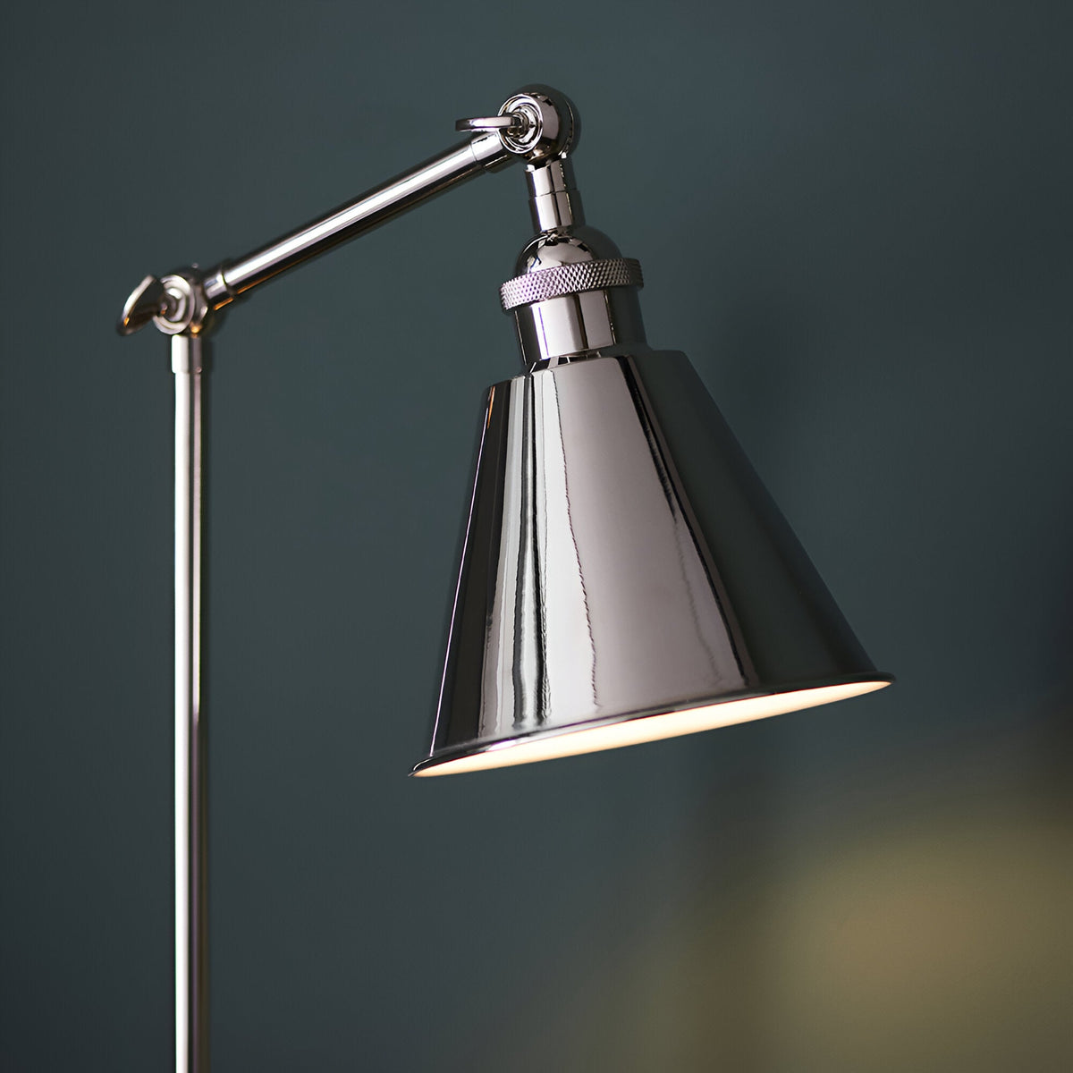 Endon Brackley table lamp in bright nickel with knurled detailing, adjustable arm and conical shade, lit against a dark teal background.