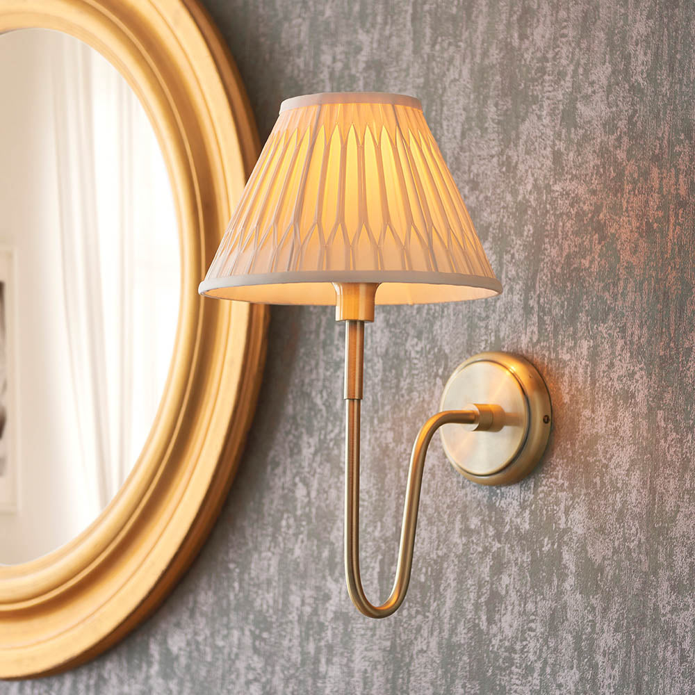 Endon Cliffdon wall light in antique brass with a curved arm and tapered pleated fabric shade, mounted beside a gold oval mirror on a grey wall.