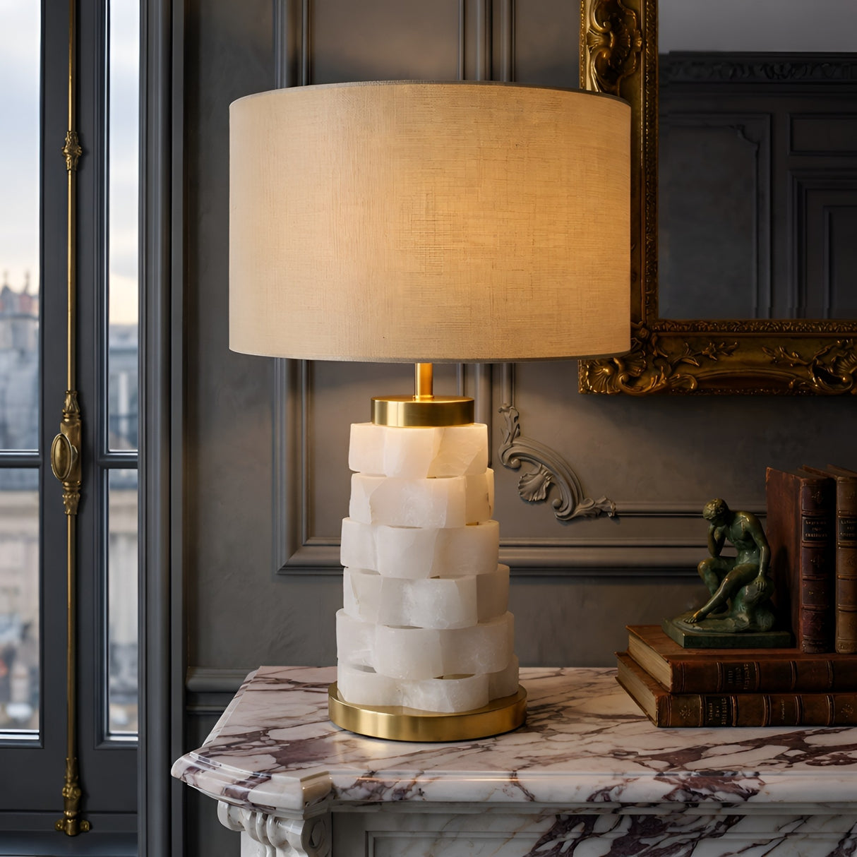 The Solmira 1 Light Stacked Alabaster Table Lamp in Brass & Soft White sits with a beige shade on a marble mantel, beside books and a green statue, below an ornate mirror and tall window in an elegant, softly lit room.