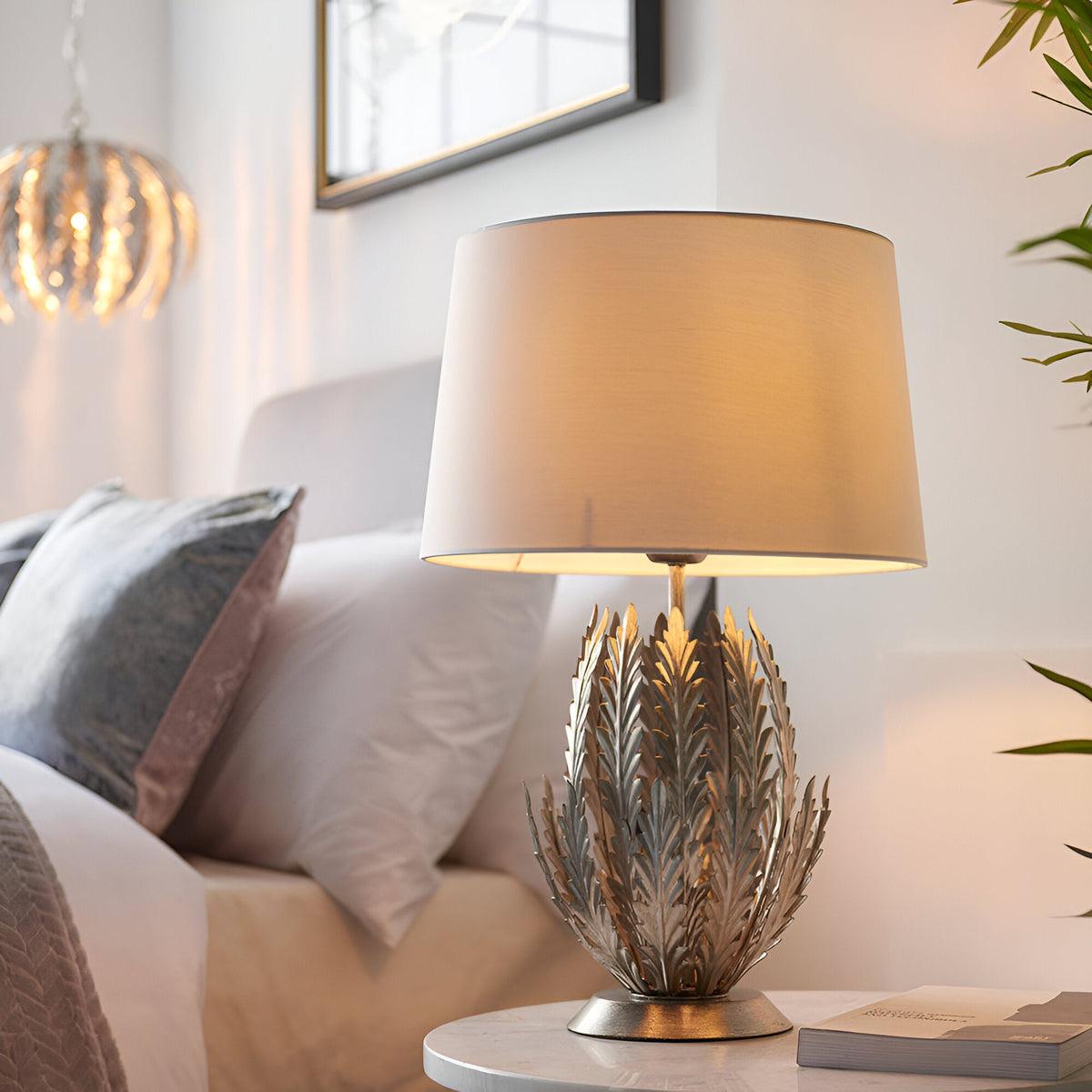 Endon Colette table lamp with layered silver leaf metal base and ivory drum shade, lit on a bedside table beside cushions and a framed mirror.