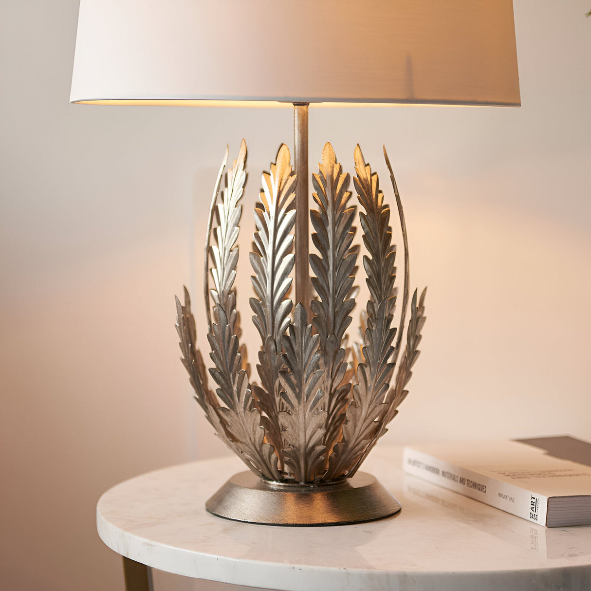 Endon Colette table lamp with layered silver leaf metal base and ivory cotton shade, lit on a round marble side table beside a book.