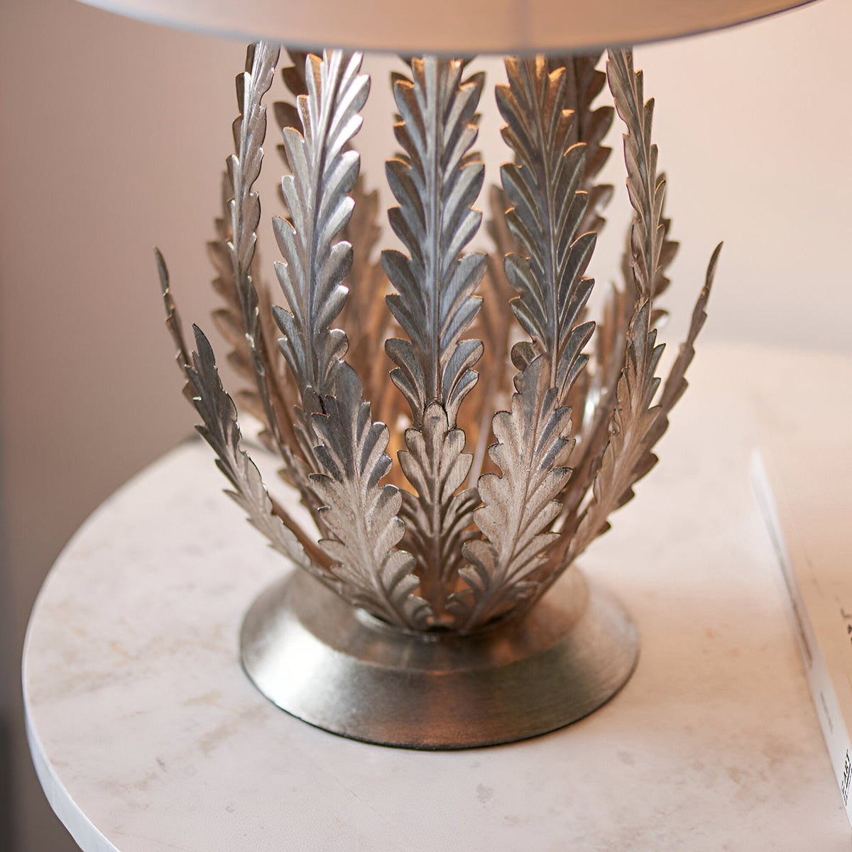 Close-up of the Endon Colette table lamp base with layered silver metal leaves on a round plinth, set on a pale side table beneath an ivory shade.